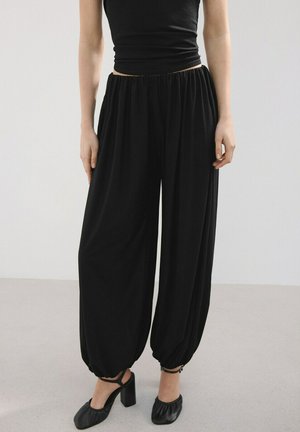 Žena nosiaca voľné čierne nohavice stiahnuté pri členkoch, čierny bezrukávový top a čierne topánky na hrubom podpätku s remienkami okolo členkov, stojaca na šedej podlahe.