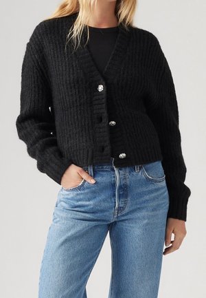 Kvinde iført sort ribstrikket cardigan med dekorative knapper og lyseblå højtaljede jeans, hånd i lommen, foran neutral baggrund.