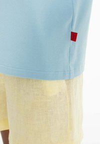 Svetle modré bavlněné tričko s krátkým rukávem, se červenou štítkovou značkou; spárováno s průsvitnými žlutými lněnými šortkami. Viditelné detaily texturované tkaniny.
