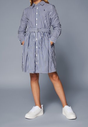 Pige iført en blå og hvid skjortekjole med lodrette striber og bælte, lange ærmer og hvide sneakers, stående med hænderne i lommerne.