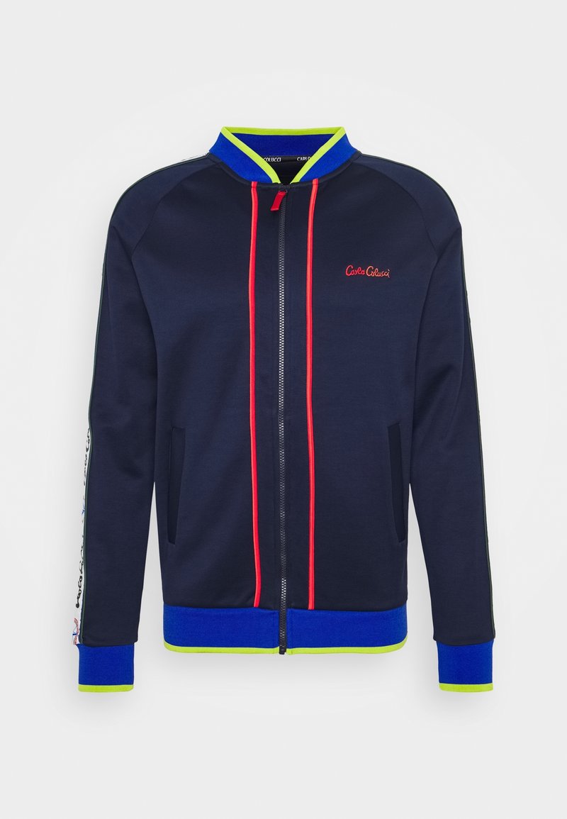 Veste zippée bleu marine avec passepoil rouge, col côtelé bleu et vert néon, poignets, taille, poches latérales et logo sur la poitrine.