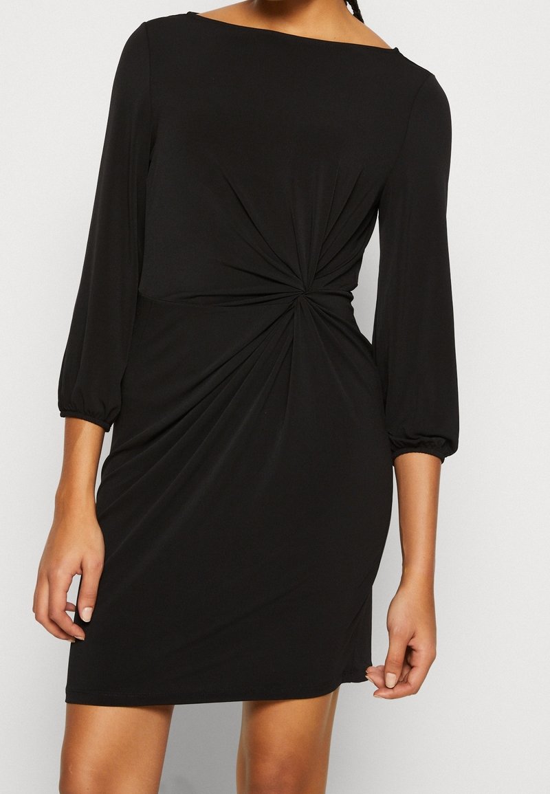 Robe noire longueur genou avec des manches trois-quarts et un détail de nœud torsadé à la taille, portée par une personne vue du cou jusqu'à mi-cuisse.