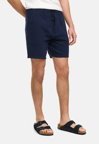 Marineblauwe shorts van textuurstof, met een elastische tailleband en een trekkoord. Gecombineerd met zwarte sandalen met twee banden en een neutraal bovenstuk.