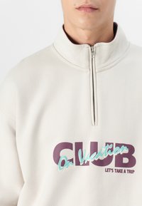 Šviesiai smėlinis zip-up džemperis su aukštu apykakle. Ant krūtinės išsiuvinėtas tekstas "CLUB On Vacation" violetine ir mėlyna spalvomis. Minkšta tekstūra.