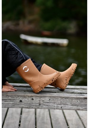 Brown rubber rain boots with a white logo, featuring a textured sole and slight mud splatter, placed on a wooden dock near water.