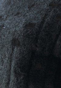 Tissu tricoté gris foncé avec des mailles visibles et de petits trous dispersés, montrant une texture complexe et des détails de fibres.