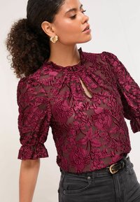 Bordová vyšívaná blúzka z priesvitnej tkaniny, s naberanými rukávmi, výstrihom na kľúč a kvetinovými vzormi. Kombinovaná s tmavými riflemi s vysokým pásom.