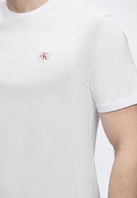 Personne portant un t-shirt blanc uni à manches courtes avec un petit logo Calvin Klein sur la poitrine gauche.