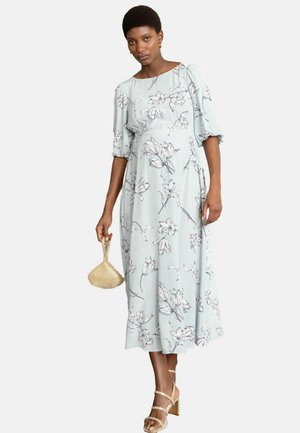 Kvinde iført en lyseblå blomsterprintet midikjole med pufærmer, der holder en gylden flettet håndtaske og har beige sandaler med stropper på.