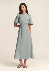Abito lungo e aderente di colore verde tenue. Presenta maniche a sbuffo corte, scollatura rotonda e dettagli arricciati in vita. Indossato con sandali minimalisti.