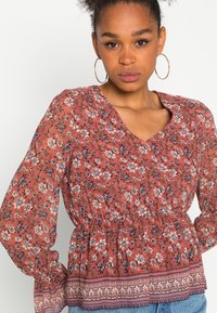 Blusa de padrão floral em terracota com decote em V, cintura elástica e mangas bufantes. Apresenta uma bainha decorativa com acabamento contrastante.
