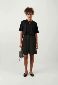 Svart avkortad t-shirt, högmidjade mörkgröna shorts och svarta platta skor. Håller en liten mönstrad väska. Enkel design med en avslappnad passform.