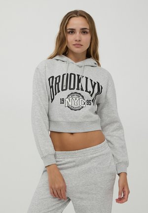 Felpa corta grigio chiaro con stampa grafica nera "BROOKLYN" e NYC. Caratterizzata da cappuccio con cordino e orlo a costine, abbinata a pantaloni della tuta coordinati.