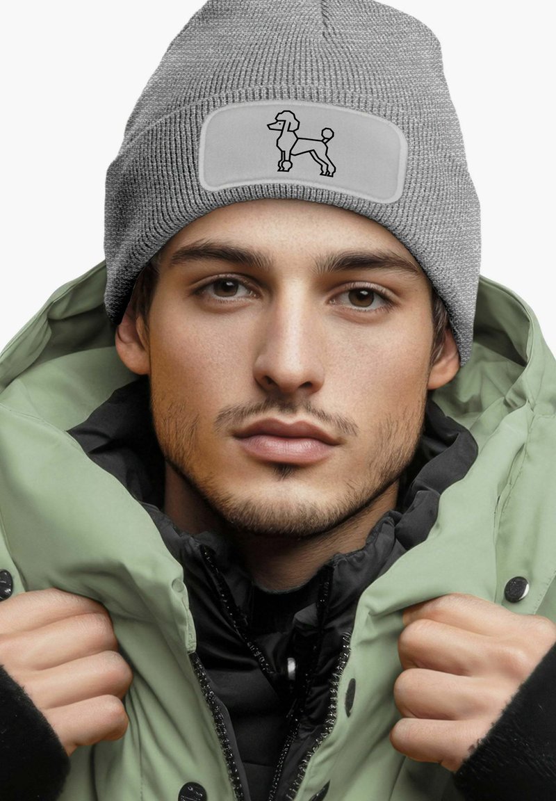 Gray knit beanie with a rectangular patch featuring a black poodle design. Worn with a green padded jacket. Close-up view of face.