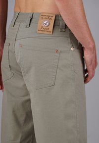 Khakifarbene Jeans mit strukturiertem Stoff, sichtbaren Nähdetails und kupferfarbenen Beschlägen. Lederetikett mit Markennamen am Bund.