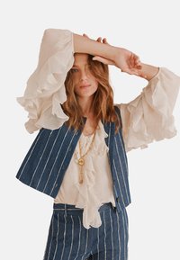 Gilet in denim con righe bianche sopra una blusa cremisi con volant. Maniche lunghe e fluenti aggiungono texture. Collana con pendente dorato in evidenza.