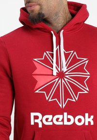 Sudadera roja con un cordón blanco y un gran logo de Reebok en blanco. Presenta un diseño gráfico que asemeja un estallido en blanco y rojo.