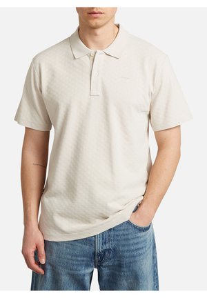 Man draagt een lichtbeige poloshirt met korte mouwen en een subtiel gestructureerd patroon, en blauwe spijkerbroek, één hand in de zak, op een witte achtergrond.