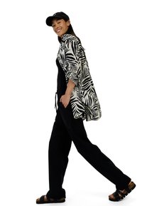 Zwart-wit shirt met bladpatroon, zwarte tanktop, losse zwarte broek en zwarte sandalen met kurken zolen. Model die loopt en glimlacht.