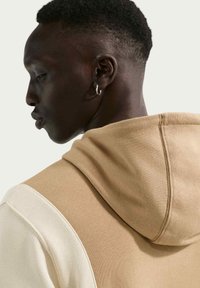 Detalle de una sudadera con capucha con un diseño en bloques de color beige y crema. Tejido suave y texturizado con una capucha prominente y costuras en las costuras laterales.