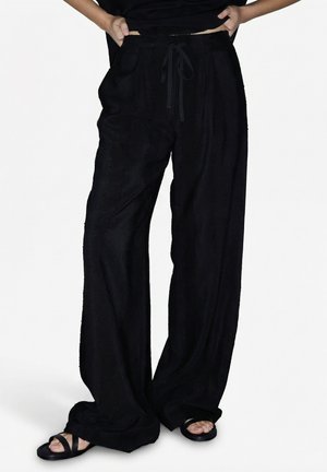 Personne portant un pantalon noir ample à cordon avec de grandes poches et des sandales noires à bout ouvert, les mains posées sur les hanches.