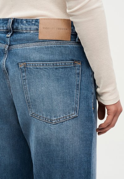 Πίσω όψη ατόμου που φοράει μπλε τζιν από τζιν υλικό με δερμάτινη ετικέτα «Tiger of Sweden» και μπεζ μακρυμάνικο μπλουζάκι.