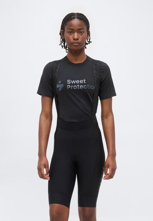 Žena s vrkočmi, ktorá má na sebe čierne cyklistické kraťasy Sweet Protection s trakmi a zodpovedajúce krátke tričko s krátkym rukávom, stojí tvárou dopredu na jednoduchom pozadí.