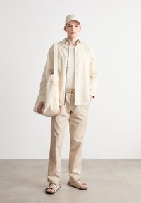 Beige bomullskjorte med knapper, matchende bukser, uformelle sandaler og en tote bag; minimalistisk design med avslappet passform og nøytrale toner.