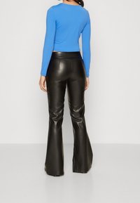 Kvinde iført en blå langærmet top og sorte højtaljede, vidde læderbukser, stående foran en ensartet lys baggrund.