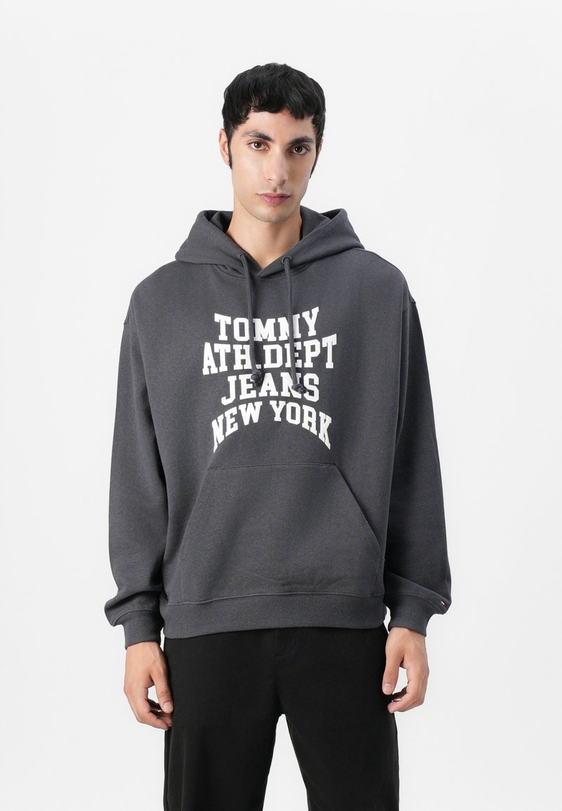 Pilkas džemperis su kapisonu, pagamintas iš minkštos medžiagos, su dideliu baltu spausdinimu: "TOMMY ATH DEPT JEANS NEW YORK." Yra priekinė kišenė ir raišteliai.