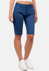 Pantaloni bermuda in denim blu con un design aderente, dotati di tasche frontali, chiusura a bottone e orli arrotolati, abbinati a sneakers bianche.