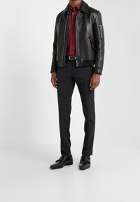 Chaqueta bomber de cuero negra con bolsillos con cremallera, camisa burdeos, pantalones negros y zapatos de hebilla negros, de pie sobre un fondo blanco.