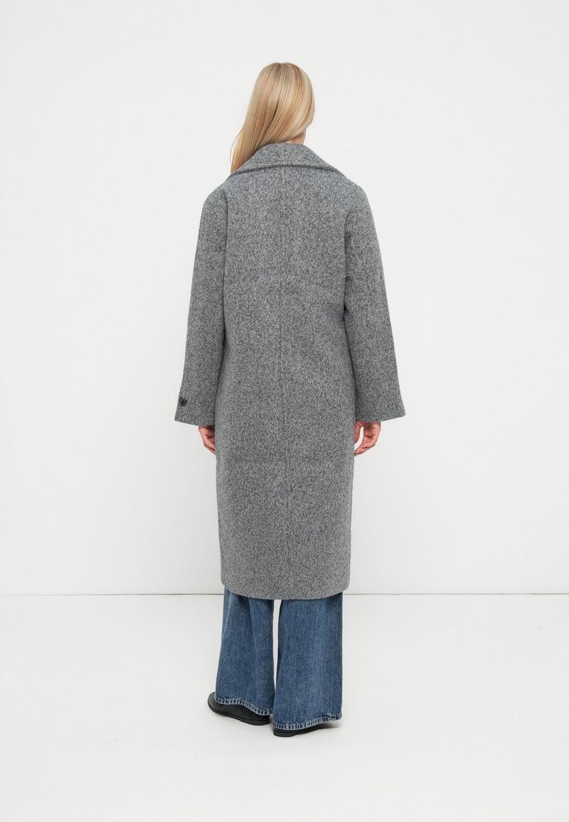 Manteau long en laine gris avec un large col, coupe ample et longueur mi-mollet. Tissu texturé avec deux boutons sur les manches, associé à un jean évasé.