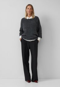Femme debout les mains dans les poches, portant un pull oversized gris foncé, une chemise blanche à col, un pantalon noir à jambes larges et des chaussures rouges à talons pointus.