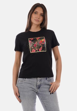 Donna che indossa una maglietta nera a maniche corte con un motivo astratto colorato a quadrati, abbinata a jeans azzurri, in posa con una mano in tasca.