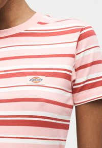 Κοντινό πλάνο ενός ατόμου που φοράει κοντομάνικο πουκάμισο με ροζ, κόκκινες και λευκές ρίγες και μικρό λογότυπο Dickies στο στήθος.