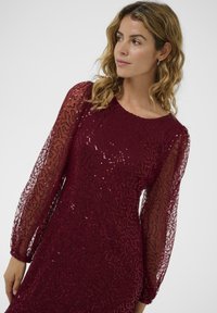 Rochie burgundy cu maneci lungi transparente și texturate, având un overlay cu un model și paiete pentru un strălucire subtilă. Decupaj rotund la gât și design ajustat.