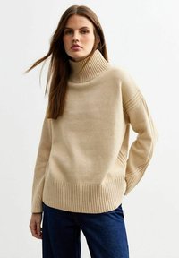 Jersey de cuello alto beige hecho de un tejido de punto suave, con dobladillo y mangas acanaladas y un sutil detalle de líneas verticales. Combinado con jeans oscuros.