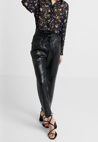 Pantaloni in pelle nera con vita alta e cintura annodata, abbinati a una blusa nera trasparente con un motivo floreale multicolore.