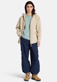 Giacca beige leggera con cappuccio, felpa blu, pantaloni cargo larghi blu navy e stivali da trekking color terra, con cerniera e polsini elastici.