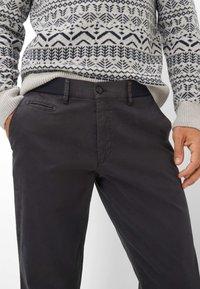 Pantaloni grigi scuri in tessuto liscio con tasche laterali e chiusura a bottone. Abbinati a un maglione a fantasia con un design geometrico.