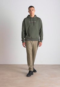 Felpa verde con cappuccio, tasca frontale e pantaloni beige, abbinati a sneakers nere. Tessuto morbido, vestibilità comoda, branding minimale.
