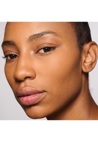 Zbliżenie twarzy z gładką skórą, neutralnym beżowym odcieniem, wyraźnie zaznaczonymi brwiami oraz naturalnie ukształtowanymi ustami. Tło w jasnoszarym kolorze.