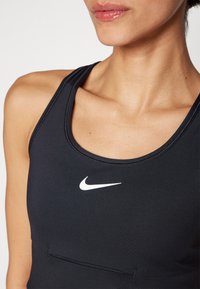 Juodas sportinis liemenukas su apvaliu kaklo iškirpimu, viduryje esantis baltas Nike logotipas. Lygi tekstūra ir prigludęs dizainas.