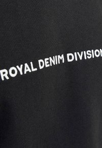 Tessuto nero con "ROYAL DENIM DIVISION" ricamato in bianco. Il tessuto è liscio con una leggera texture, che enfatizza il testo in modo evidente.
