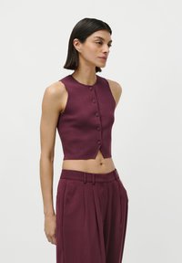 Žena s rovnými tmavými vlasmi, oblečená v bezrúchom skrátenom košelovom vrchu a vysoko pásových širokých nohaviciach v burgundskej farbe.