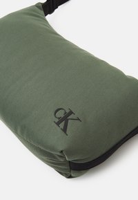 Borsa a tracolla verde in tessuto con una superficie liscia, caratterizzata da un logo nero e una tracolla nera a contrasto. Forma rettangolare con bordi arrotondati.