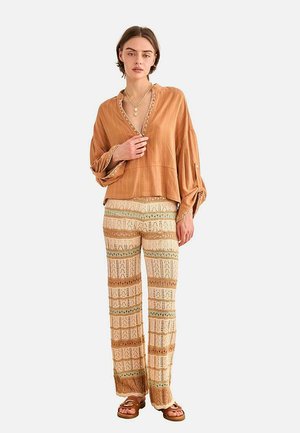 Mujer con blusa marrón suelta, pantalones de ganchillo en crema y marrón con estampado, sandalias marrones y un collar de varias capas sobre un fondo blanco.