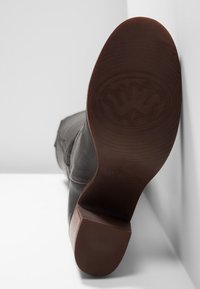 Vue de la semelle d'une botte en cuir noir avec un talon marron, posée à la verticale contre un mur gris clair et un sol blanc.