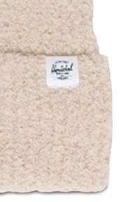 Tessuto beige soffice con una superficie testurizzata. Presenta un'etichetta bianca con testo nero, che indica il nome del marchio o del prodotto.